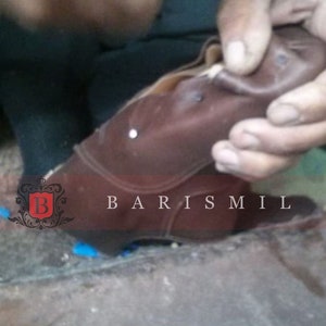 May include: A close-up of a brown leather boot being worked on by a shoemaker. The boot has a white stitch detail and the text "BARIS MIL" is visible on the side.
