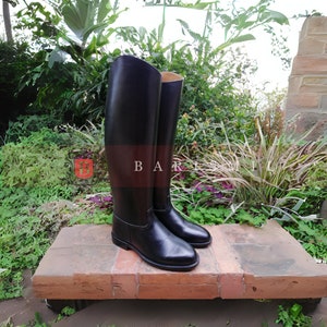 Puede incluir: Un par de botas de montar altas de cuero negro. Las botas se exhiben al aire libre sobre una superficie de ladrillo, con exuberante vegetaci&oacute;n en el fondo. El texto "BAR" y "MILL" es visible.