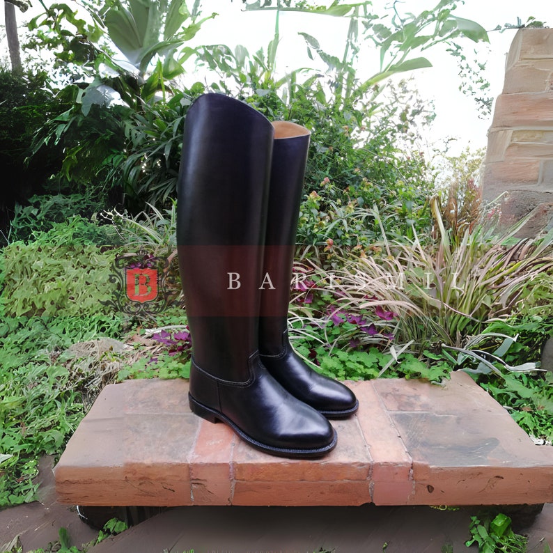 Puede incluir: Un par de botas de montar altas de cuero negro. Las botas se exhiben al aire libre sobre una superficie de ladrillo, con exuberante vegetaci&oacute;n en el fondo. El texto "BAR" y "MILL" es visible.