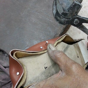 May include: A close-up of a brown leather shoe being stitched together with a metal tool. The shoe is being held in place by a hand.