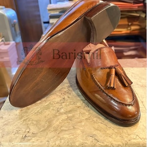 May include: A pair of brown leather tassel loafers with a brown leather sole. The loafers have a tassel detail on the front and a stitched design around the toe.
