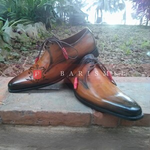 May include: A pair of brown leather dress shoes with black leather toes. The shoes have laces and red accents on the laces. The shoes are on a brick surface.