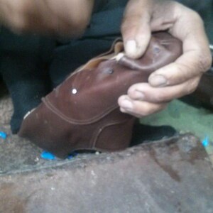 May include: A person is working on a brown leather shoe. The shoe is being held in their hand and they are using a tool to work on it. There are small metal pieces scattered on the surface.