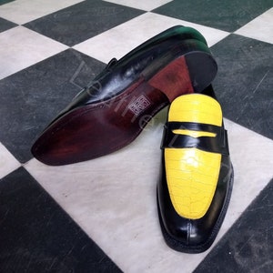 May include: A pair of black leather loafers with a yellow crocodile-embossed leather panel on the toe. The loafers have a brown leather sole and a black leather upper.