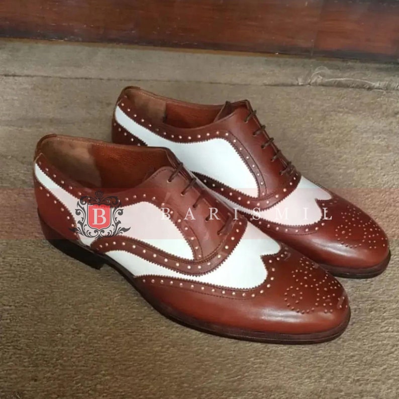 May include: A pair of brown leather brogue shoes with white leather accents. The shoes have a lace-up closure and a wingtip design. The shoes are made of high-quality leather and have a classic look.