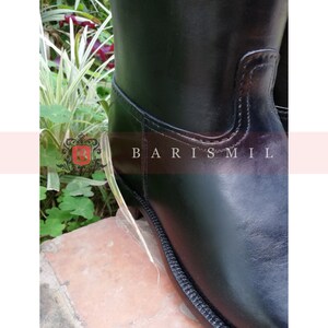 Puede incluir: Primer plano de una bota de montar de cuero negro. La bota presenta un acabado liso y brillante y un detalle de costura en la suela. El texto "BARIS MIL" es visible en el lateral. La bota se encuentra sobre un fondo de vegetaci&oacute;n.