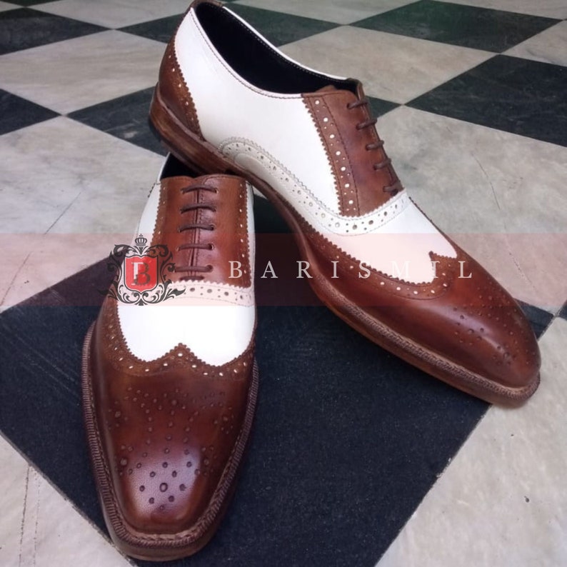 May include: A pair of brown and white leather brogue shoes with a wingtip design. The shoes have a lace-up closure and a brown leather sole.