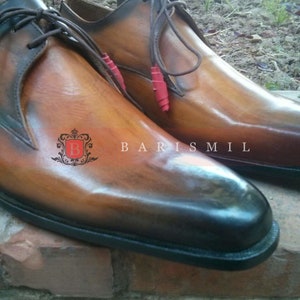 May include: A pair of brown leather dress shoes with black laces and a black sole. The shoes have a distressed look and are made of high-quality leather. The brand name "BARISMIL" is visible on the shoes.