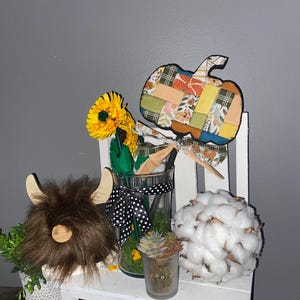 May include: A decorative arrangement featuring a brown faux fur highland cow head, a glass vase with yellow sunflowers and a black and white polka dot ribbon, a fabric pumpkin on a stick, a small succulent, and a white cotton ball sphere.