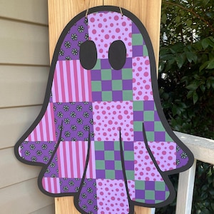 May include: A patchwork ghost-shaped wooden sign with a black outline. The ghost features various patterns in shades of purple, pink, and green, including stripes, polka dots, and floral designs. Two black oval cutouts serve as eyes. A jute string is attached for hanging.