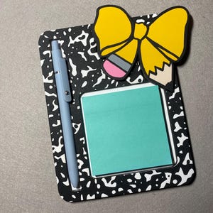 Op de afbeelding: Een notitieblok met een zwart-wit gespikkeld ontwerp, een lichtblauwe pen en een gele potloodstrik. In het midden zit een turquoise plakbriefje. De potloodstrik heeft een roze gum en een beige potloodpunt.