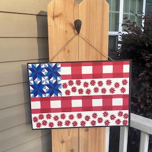 Puede incluir: Un letrero rectangular de madera con temática patriótica. El diseño presenta un patrón similar a una bandera con formas de estrellas azules, rayas a cuadros rojas y blancas y detalles de flores rojas. El letrero está colgado de un poste de madera.