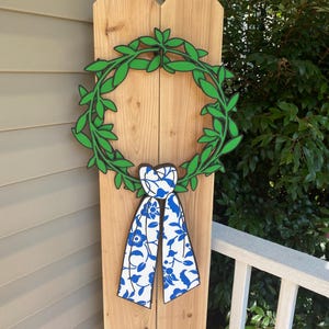 May include: A wooden wreath with a green leaf design and a blue and white floral bow. The wreath is mounted on a wooden post. The wreath is a decorative item.