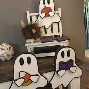 May include: Wooden Halloween decorations featuring white ghost figures with black eyes. One ghost holds a pumpkin, another a piece of candy corn, and the third a book. A Highland cow figurine and small purple rectangles are also present.