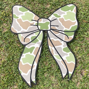 May include: A large, green, brown, and white camouflage patterned bow. The bow is made of a thick, sturdy material and has a glossy finish.
