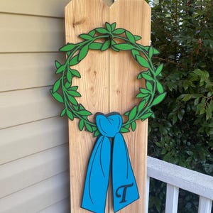 May include: Wooden sign featuring a green wreath with black outlines and a blue ribbon. The ribbon has the letter "T" on it. The sign is made of light brown wood and is displayed outdoors.