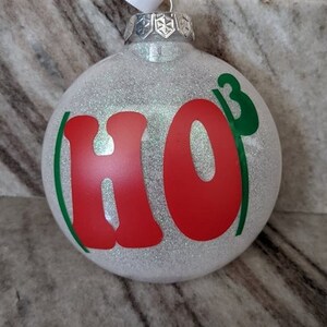 May include: White glitter Christmas bauble with the word "HO" in red and a green "3" to the right of the "O".