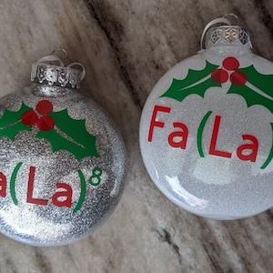 May include: Two silver glitter Christmas baubles with green holly leaves and red berries. The baubles have the text "Fa(La)8" on them.