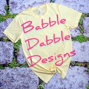 May include: A yellow t-shirt with the words "Babble Drabble Designs" written in pink on it.