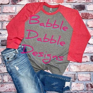 May include: A gray and red raglan shirt with the text "Babble Dabble Designs" in pink, paired with blue denim jeans. The jeans have distressed details.