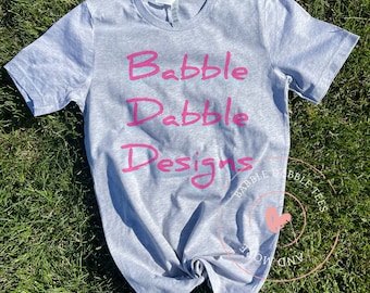 Könnte beinhalten: Ein hellgraues T-Shirt mit dem Text "Babble Dable Designs" in Pink mit blauem Umriss.