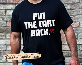 Könnte beinhalten: Ein schwarzes T-Shirt mit weißem Text, der "PUT THE CART BACK." lautet. Neben dem Text befindet sich ein rotes Einkaufswagen-Symbol. Das Shirt wird von einer Person getragen.