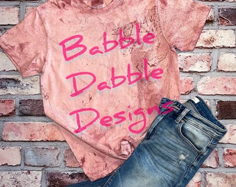 Könnte beinhalten: Ein pinkfarbenes T-Shirt mit dem Schriftzug "Babbble Dabble Designs" in Pink. Das Shirt liegt auf einer Backsteinmauer mit einer blauen Jeans.