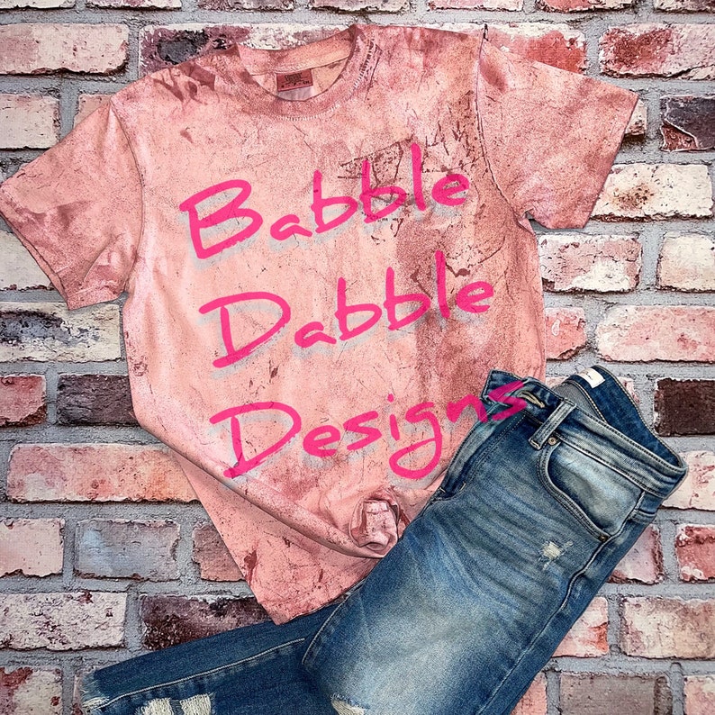 May include: A pink t-shirt with the words "Babbble Dabble Designs" written in pink on it. The shirt is laying on a brick wall with a pair of blue jeans.