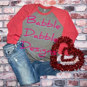 Könnte beinhalten: Ein Baseball-T-Shirt in Grau meliert und Korallenrot mit dem Text "Babble Drabble Designs" in pinkfarbenen Buchstaben. Das Shirt liegt auf einem Ziegelhintergrund mit einer roten herzförmigen Girlande.