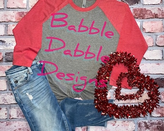 Könnte beinhalten: Ein Baseball-T-Shirt in Grau meliert und Korallenrot mit dem Text "Babble Drabble Designs" in pinkfarbenen Buchstaben. Das Shirt liegt auf einem Ziegelhintergrund mit einer roten herzförmigen Girlande.