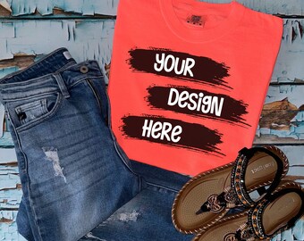 Könnte beinhalten: Korallfarbenes T-Shirt mit dem weißen Text "YOUR DESIGN HERE", kombiniert mit blauen Jeans und braunen Sandalen. Die Jeans haben Distressed-Details. Die Sandalen sind mit dekorativen Perlen verziert.