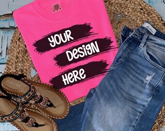 Könnte beinhalten: Ein leuchtend pinkfarbenes T-Shirt mit schwarzen Pinselstrichen und dem Text "YOUR DESIGN HERE". Das Shirt liegt flach auf einer gewebten Oberfläche mit einem Paar braunen Sandalen und einer blauen Jeans.