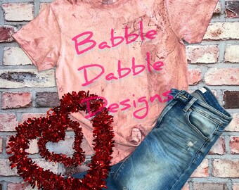 Könnte beinhalten: Ein pinkfarbenes Batik-T-Shirt mit dem Text "Babbble Dabble Designs" in pinkfarbener Schrift. Das Shirt wird mit einer blauen Jeans und einer roten Herzform-Dekoration kombiniert.