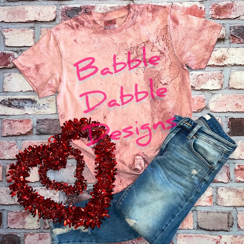 K&ouml;nnte beinhalten: Ein pinkfarbenes Batik-T-Shirt mit dem Text "Babbble Dabble Designs" in pinkfarbener Schrift. Das Shirt wird mit einer blauen Jeans und einer roten Herzform-Dekoration kombiniert.