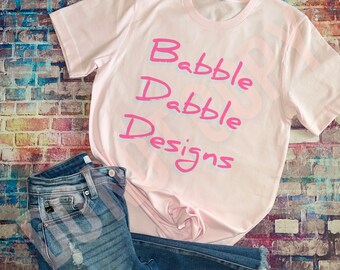 Könnte beinhalten: Ein hellrosa T-Shirt mit dem Text "Babble Dabble Designs" in leuchtendem Pink. Das Shirt wird mit blauen Jeans mit Distressed-Details kombiniert. Die Artikel sind vor einem Backsteinmauer-Hintergrund angeordnet.