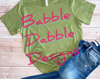 Könnte beinhalten: Ein grünes T-Shirt mit den Worten "Babble Drabble Designs" in Pink auf der Vorderseite.