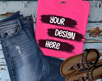 Könnte beinhalten: Ein pinkfarbenes T-Shirt mit schwarzen Pinselstrichen und dem Text "YOUR DESIGN HERE" darauf. Das T-Shirt liegt auf einer blau-weißen Holzoberfläche mit einer blauen Jeans und braunen Sandalen.