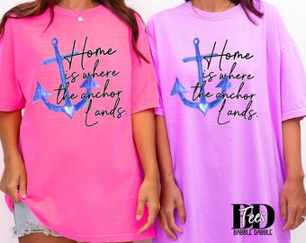 Könnte beinhalten: Zwei übergroße T-Shirts, eines in Pink und eines in Lavendel, mit einem blauen Anker-Motiv und dem Schriftzug "Home is where the anchor lands." Die Shirts haben kurze Ärmel und eine lockere Passform, ideal für Freizeitkleidung.