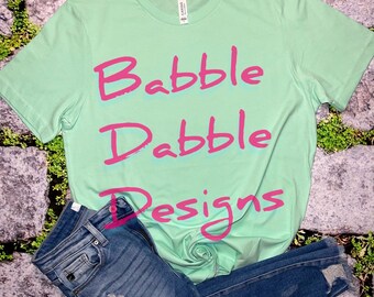Könnte beinhalten: Ein hellgrünes T-Shirt mit dem Text "Babble Drabble Designs" in rosa Schrift.