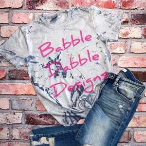Könnte beinhalten: Ein graues Batik-T-Shirt mit dem Text "Brabble Dabble Designs" in Pink.