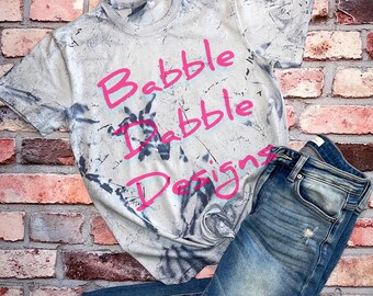 Könnte beinhalten: Ein graues Batik-T-Shirt mit dem Text "Brabble Dabble Designs" in Pink.