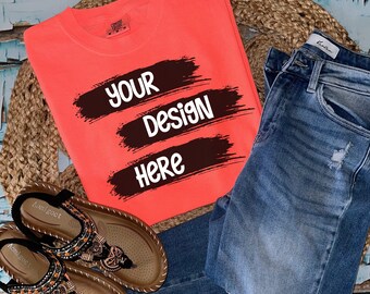 Könnte beinhalten: Ein korallenfarbenes T-Shirt mit dem Text "YOUR DESIGN HERE" in schwarzen Farbstrichen. Das T-Shirt liegt auf einer geflochtenen Strohmatte mit einem Paar braunen Sandalen und einer blauen Jeans.