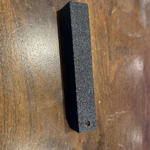 May include: A black, rectangular object with a textured surface and a small hole at one end. The object is positioned vertically against a wooden background, showcasing its geometric shape and dark color.