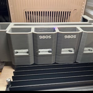 May include: A gray 3D-printed organizer with multiple compartments. The organizer has the number "9805" printed on it. The organizer is sitting on a black, ridged surface. The background includes a beige panel with vertical slats.