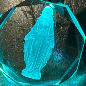 May include: A teal-colored, faceted crystal ornament featuring an etched image of a religious figure. The figure is illuminated, creating a glowing effect within the clear crystal. The ornament has a geometric shape.