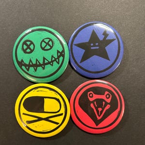 May include: Four circular pinback buttons featuring graphic designs. One is green with a stitched-mouth smiley face, one is blue with a black star, one is yellow with a pill icon, and one is red with a stylized black animal head graphic.
