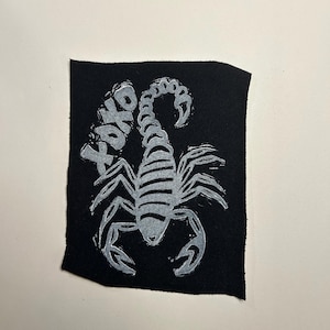 May include: A black fabric patch featuring a silver-colored screen-printed graphic of a scorpion with its tail curved upward. The text XOXO is printed in a matching silver color to the left of the scorpion, with a textured, hand-drawn appearance.
