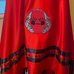May include: A red shawl with black fringe and a black ribbon trim. The ribbon has a repeating pattern of black handprints. The shawl has a large embroidered patch with two black handprints and white feathers.