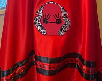 May include: A red shawl with black fringe and a black ribbon trim. The ribbon has a repeating pattern of black handprints. The shawl has a large embroidered patch with two black handprints and white feathers.