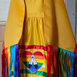 Op de afbeelding: Een gele satijnen jurk met een regenboog hartontwerp en franjes. Het hart heeft een Native American symbool en de regenboogkleuren zijn in de volgorde van de Pride-vlag.